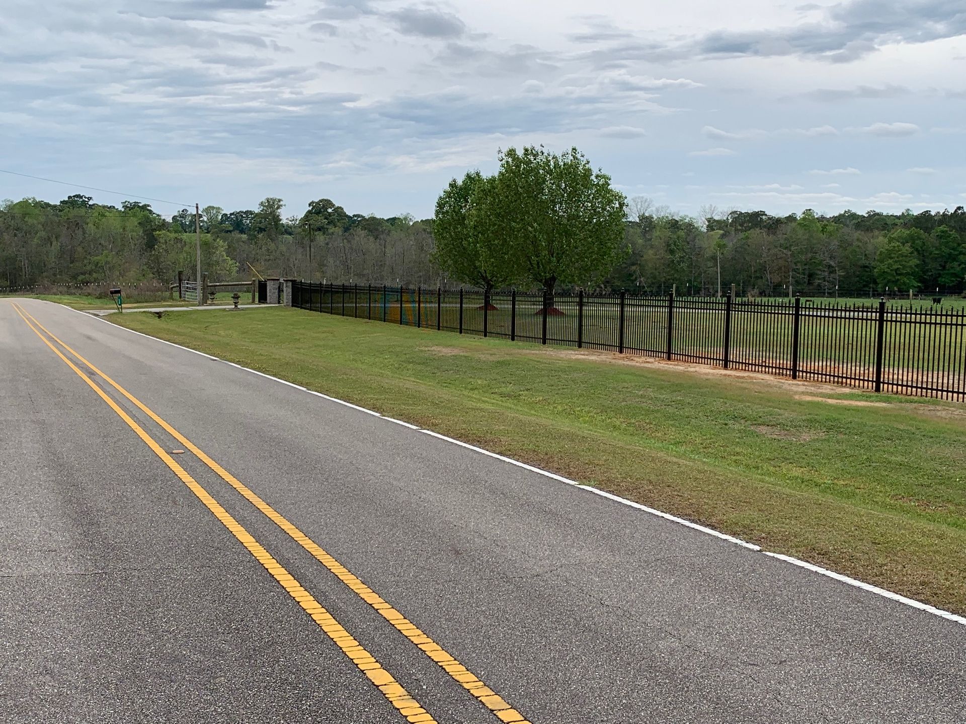 Empty rural road with double yellow lines beside a fenced grassy field and trees under a cloudy sky