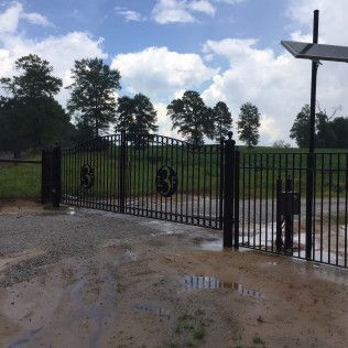 Black metal gate opening to a grassy field under a cloudy sky, with muddy puddles in the foreground