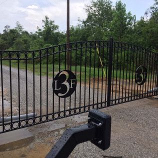 Black metal gate with “L3F” and “3” emblems along a driveway, with trees and gravel in the background