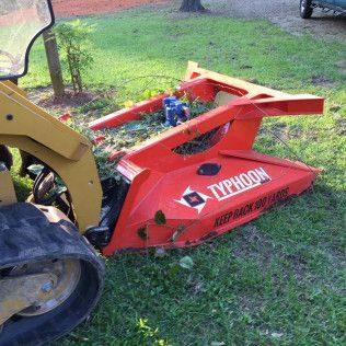 Red Typhoon skid-steer mulcher attachment in a grassy yard, connected to a loader arm.