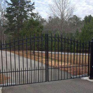 Black metal driveway gate with curved top in front of a paved road and trees
