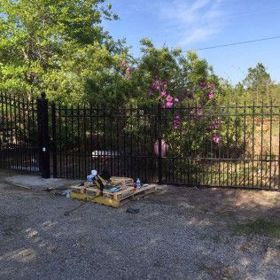 Black metal fence beside a gravel road, with trees and pink flowers behind it and stacked materials in front