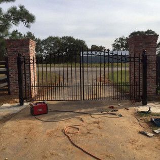Closed black iron gate between brick pillars at the end of a driveway, with a red toolbox in front.