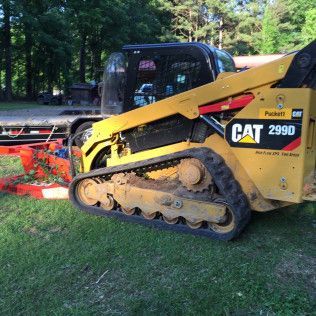 Yellow CAT 299D tracked skid steer on grass near an orange attachment, with trees in the background