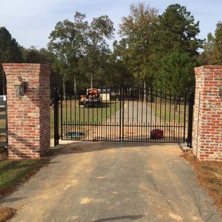 Closed black gate between brick pillars, with a driveway leading to a tree-lined yard and parked utility vehicle.