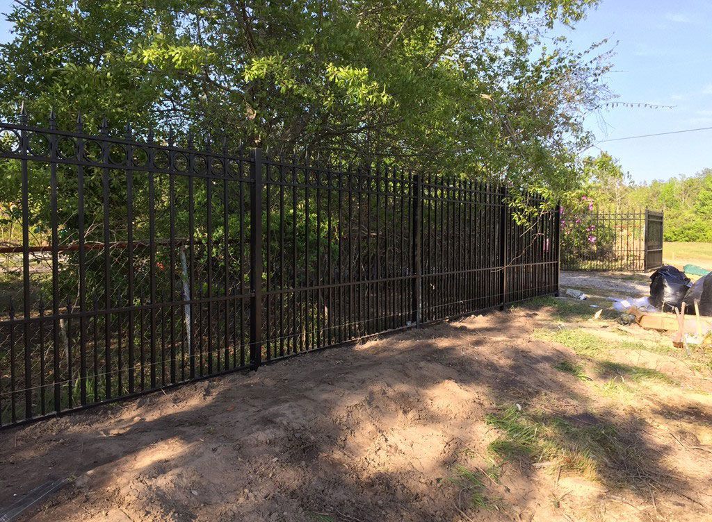 Black metal fence along a dirt path beside trees and a brick pillar in sunny daylight