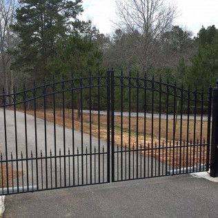 Black metal driveway gate across a paved entrance, with trees and brush behind it.