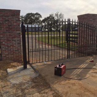 Black metal gate between brick pillars, with a red battery charger on the ground in a driveway.