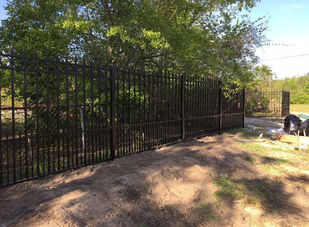 Black metal fence along a dirt path beside leafy trees in bright sunlight