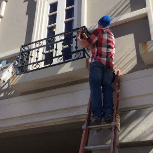 Worker on a ladder installing black metal balcony railing on a beige house exterior
