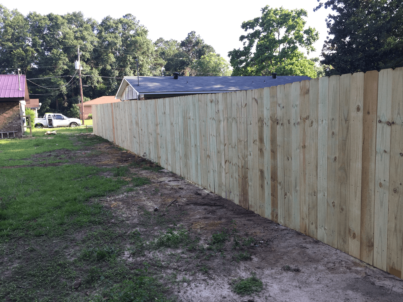 Long wooden fence along a dirt path beside grass, with trees and a small building in the background.