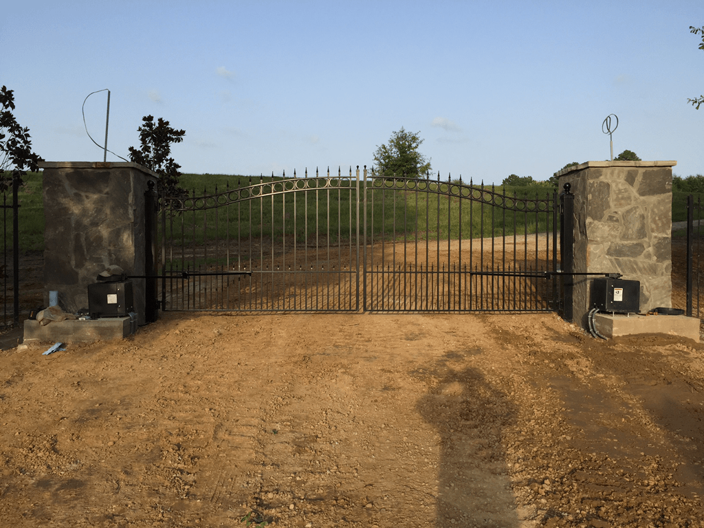 Closed black iron gate between stone pillars at the end of a dirt driveway under a clear sky
