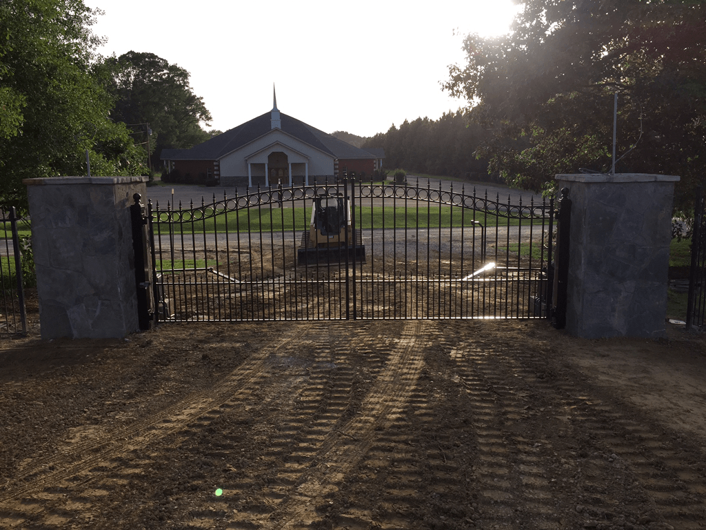Closed black iron gate in front of a small building at sunset, with a dirt driveway leading up to it