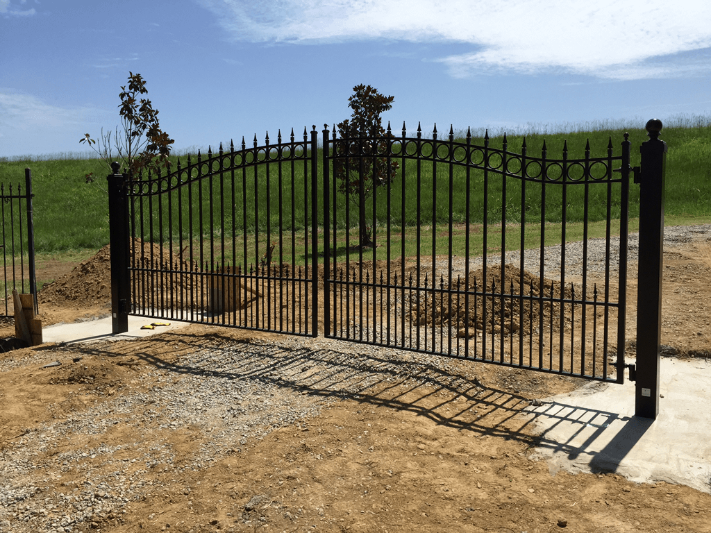 Black iron gate across a dirt driveway in front of a green field under a blue sky