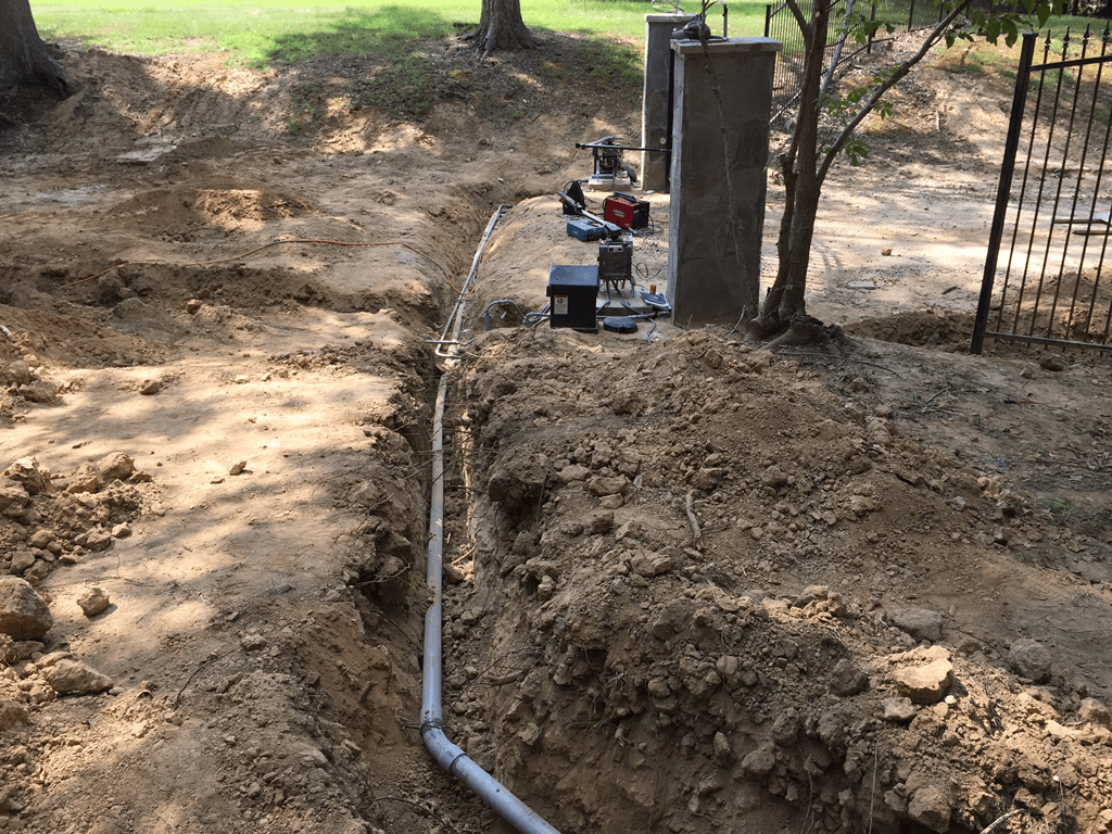 Trench with a white pipe running through dirt beside a gate and utility equipment outdoors