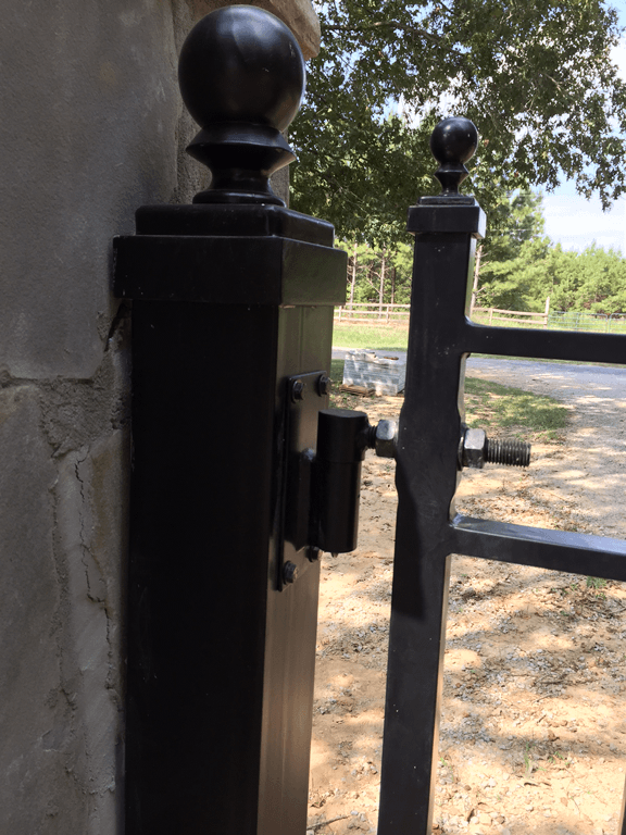 Black metal gate latch and post beside a stone wall, opening to a sunny dirt path and trees.