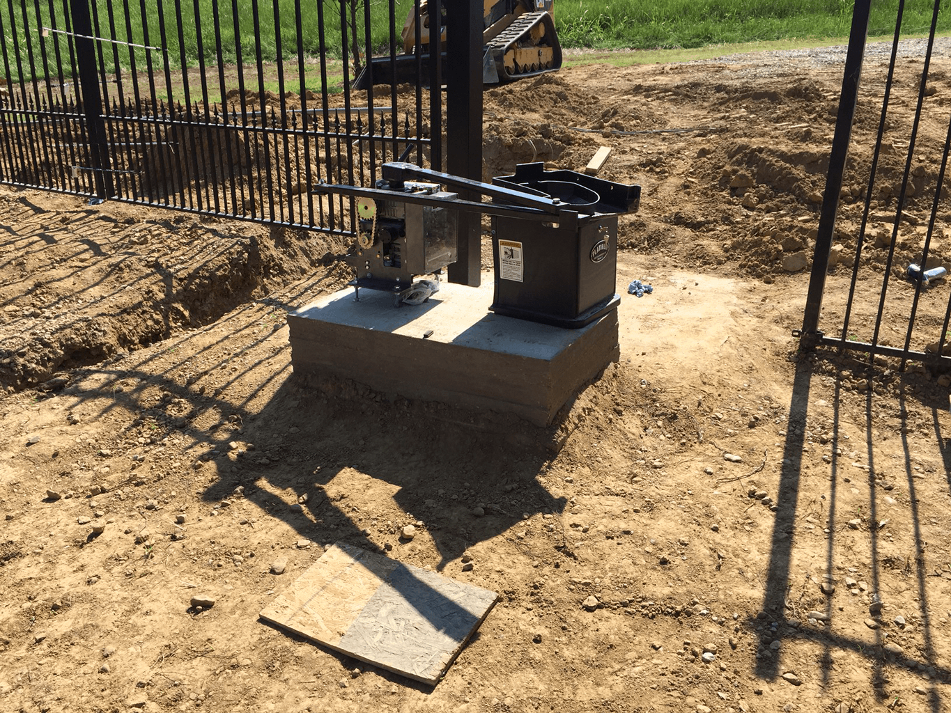 Metal utility equipment on a concrete pad beside a fenced dirt area outdoors