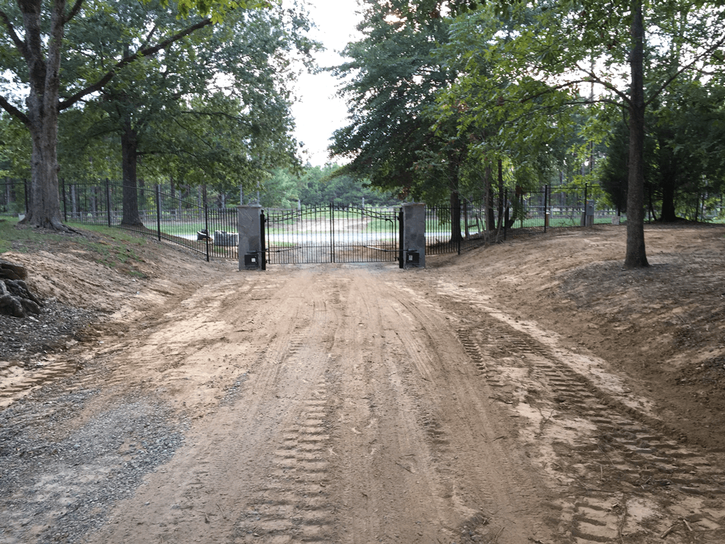 Dirt road leading to a gated park entrance, flanked by trees and grassy slopes.