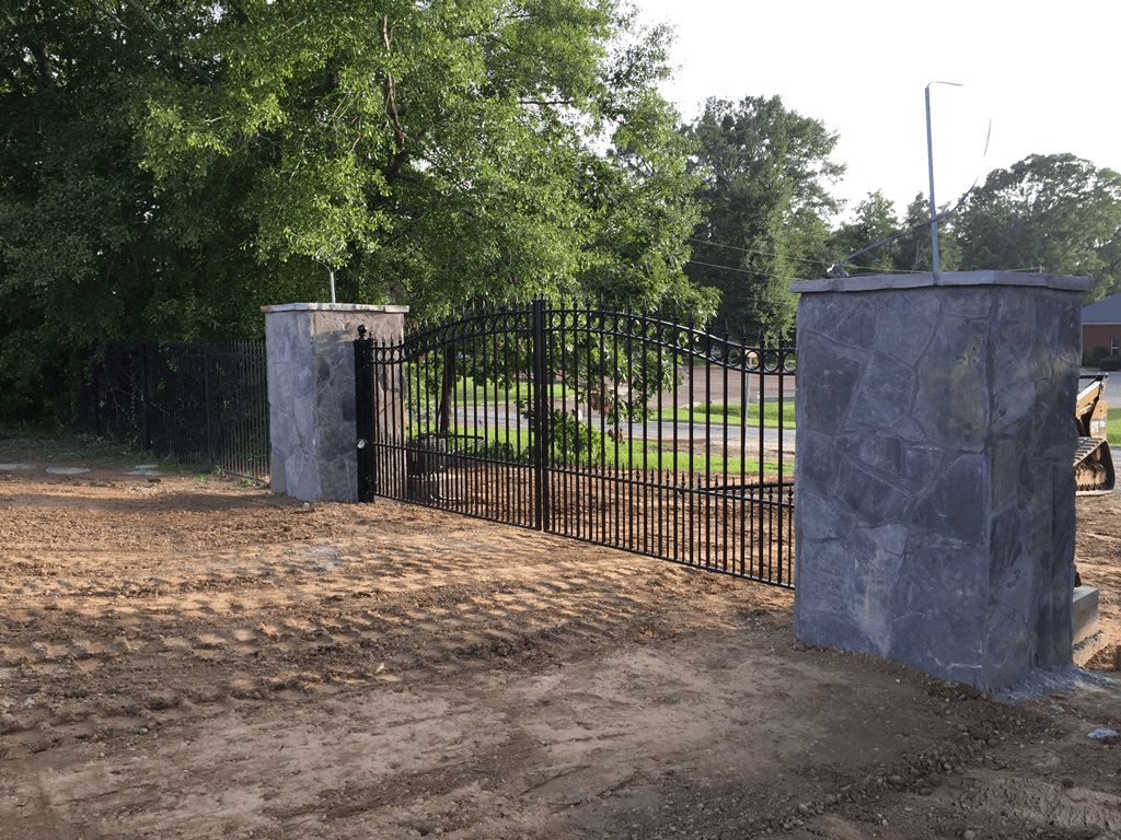 Black metal gate between stone pillars at the edge of a dirt driveway, with trees and grass behind it