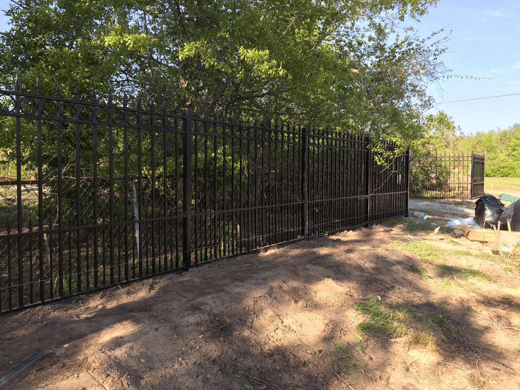 Black metal fence along a dirt path beside trees and a creek under bright sunlight