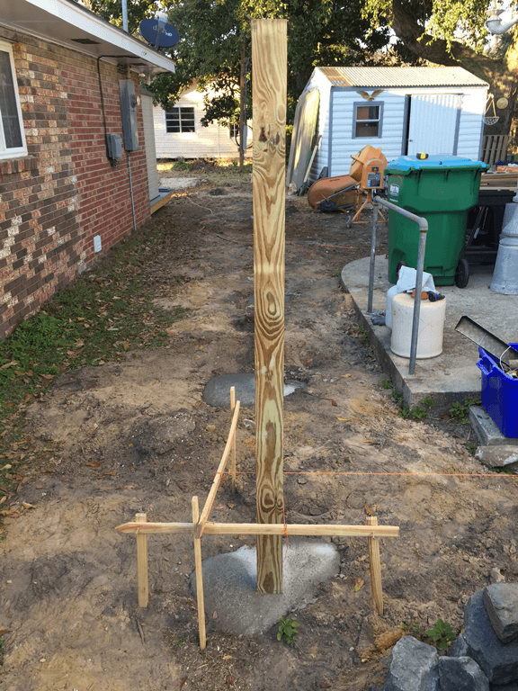 Wooden support post braced with stakes in a dirt side yard beside a brick house and trash bins