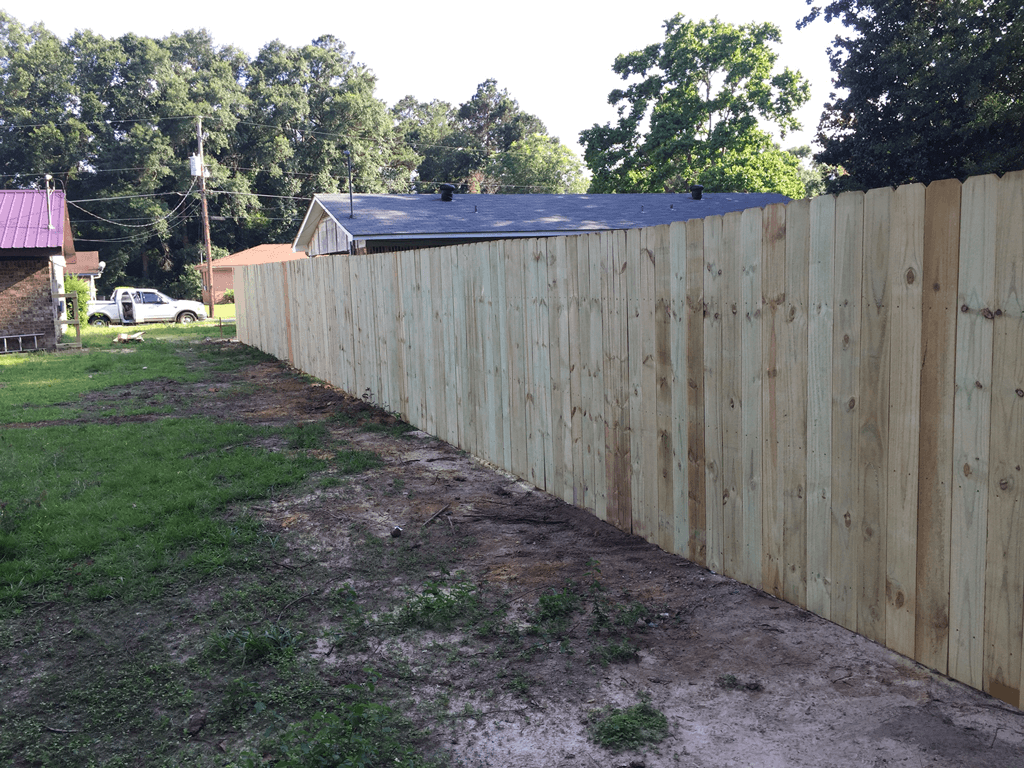 Fresh wooden privacy fence along a dirt path, with trees and a house in the background.