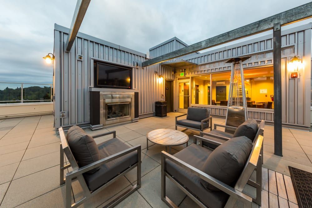 Modern rooftop patio at twilight with comfortable seating, a sleek outdoor fireplace with a mounted TV above, ambient lighting, and warm interior lights visible through glass doors at Marq West Seattle in Seattle, WA.
