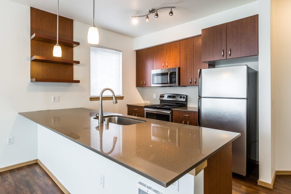 Modern kitchen interior with wooden cabinets and stainless steel appliances. A central island featuring a sink and a beige countertop extends into the foreground, with pendant lights hanging above. On the right side, there's a refrigerator next to an oven range with a microwave above it. The kitchen has a window with blinds, providing natural light, and hardwood flooring at Marq West Seattle in Seattle, WA.