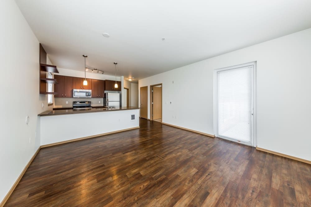 Image of a modern, empty living room with hardwood floors, an open-concept kitchen with dark cabinets and stainless steel appliances, contemporary light fixtures, and a sliding glass door with blinds leading to an unseen outdoor space at Marq West Seattle in Seattle, WA.