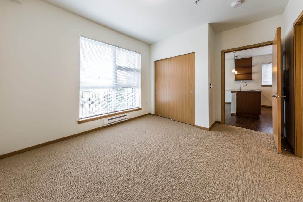 Empty bedroom in a modern apartment with beige carpeting, a large window with blinds, and an open door leading to a kitchen with wooden cabinets and a hanging pendant light fixture at Marq West Seattle in Seattle, WA.