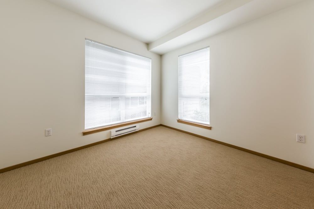 Interior image showing an empty room with beige carpet flooring, white walls, and two windows with white blinds. The room appears well-lit and is unfurnished, suggesting a blank canvas for decoration at Marq West Seattle in Seattle, WA.