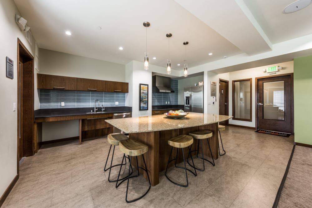 Modern clubhouse kitchen in a communal area featuring a large curved island with granite countertops and bar stools, sleek wood cabinetry with dark countertops, stainless steel appliances, and pendant lighting at Marq West Seattle in Seattle, WA.