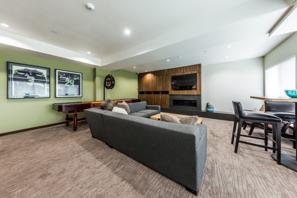 A contemporary clubhouse in neutral tones with green accent walls. The space features a gray sectional sofa adorned with pillows, facing a dark wooden entertainment center with a large flat-screen TV above an electric fireplace. To the left, there's a polished black upright piano against the wall and three framed artworks above. On the right, a high wooden bar table with two chairs is situated beside a white window with blinds. The room has recessed lighting and a brown carpeted floor at Marq West Seattle in Seattle, WA.