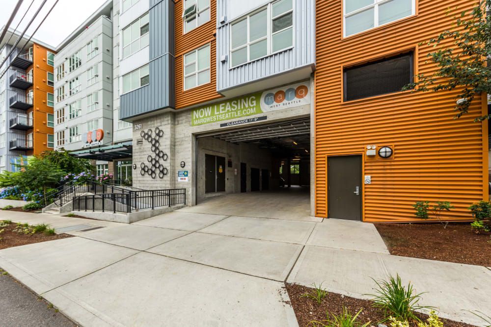 Exterior view of a modern apartment complex with a mix of gray and orange paneling, featuring a 'Now Leasing' sign and an underpass entrance to covered parking at Marq West Seattle in Seattle, WA.