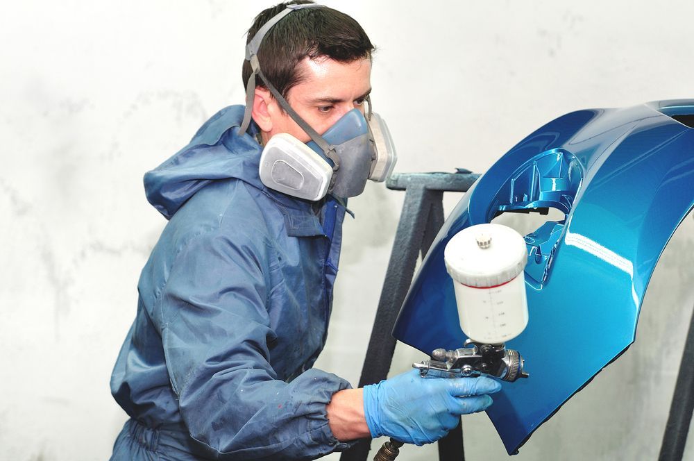 A Man Wearing a Mask and Gloves is Spray Painting a Blue Car Bumper — Lyons Street Paint Panel in Bungalow, QLD