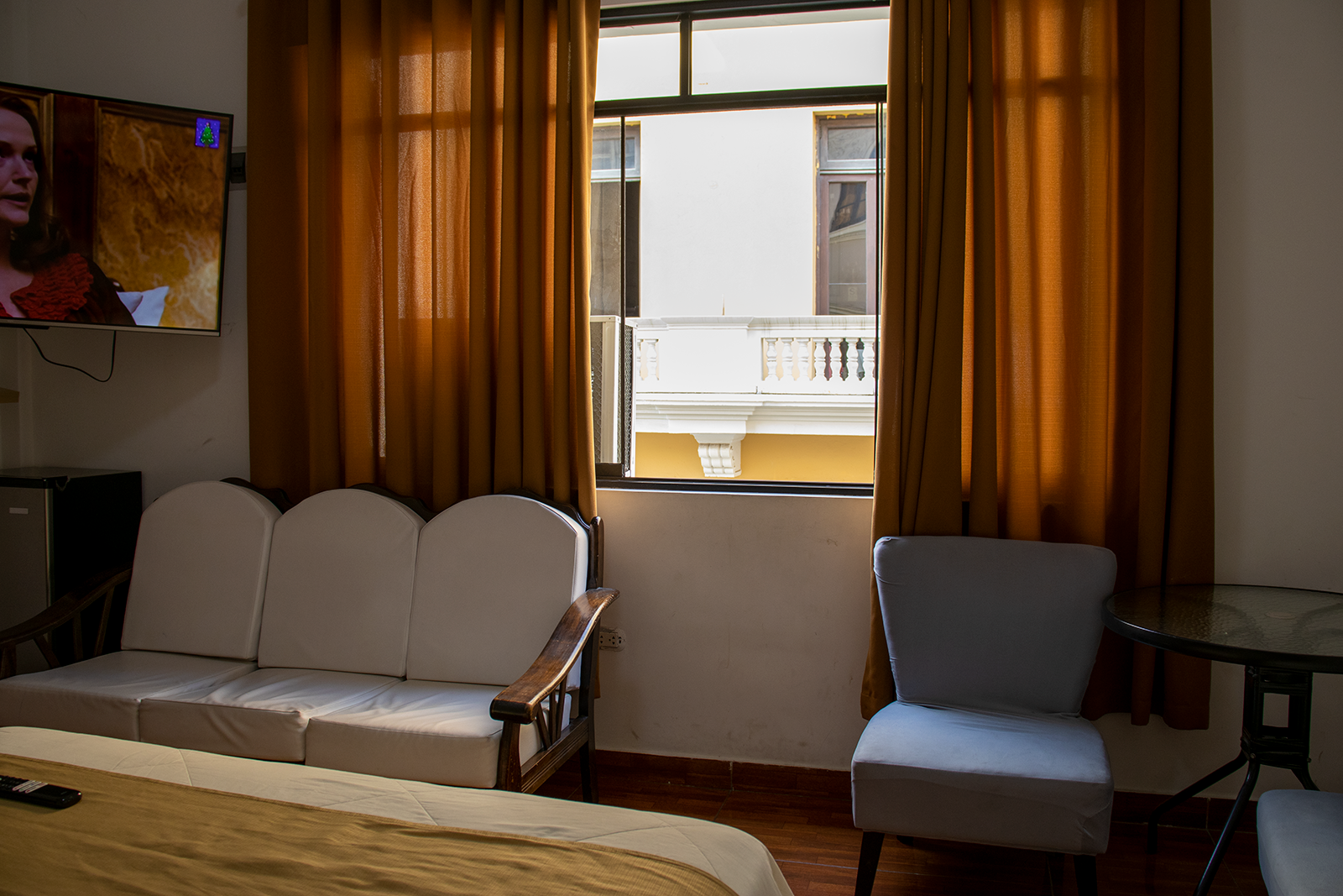 Hotel room interior: couch, chair, TV, window with balcony view, curtains, and table.