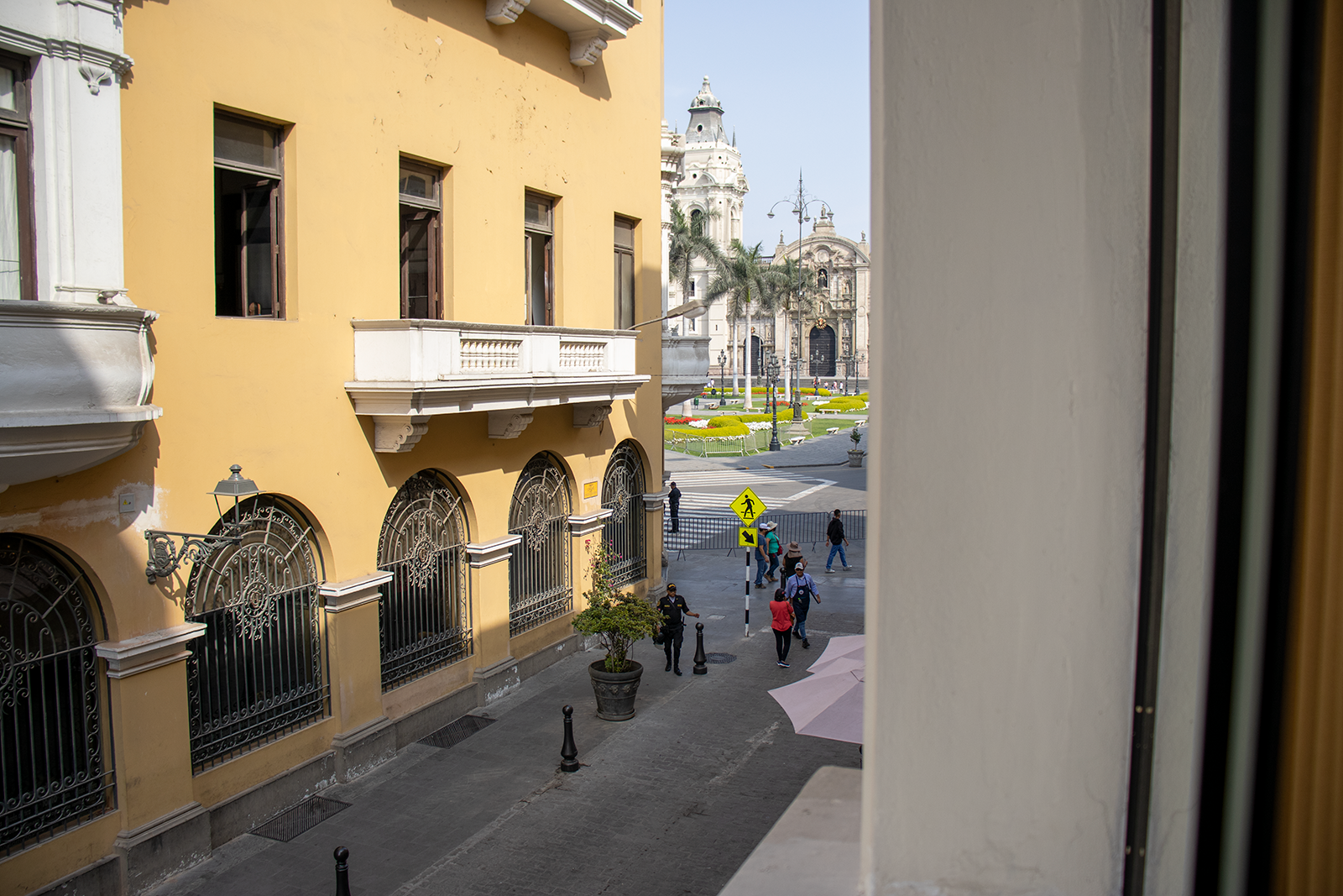 Yellow building with ornate windows overlooks a street and plaza with people and a church in the distance.