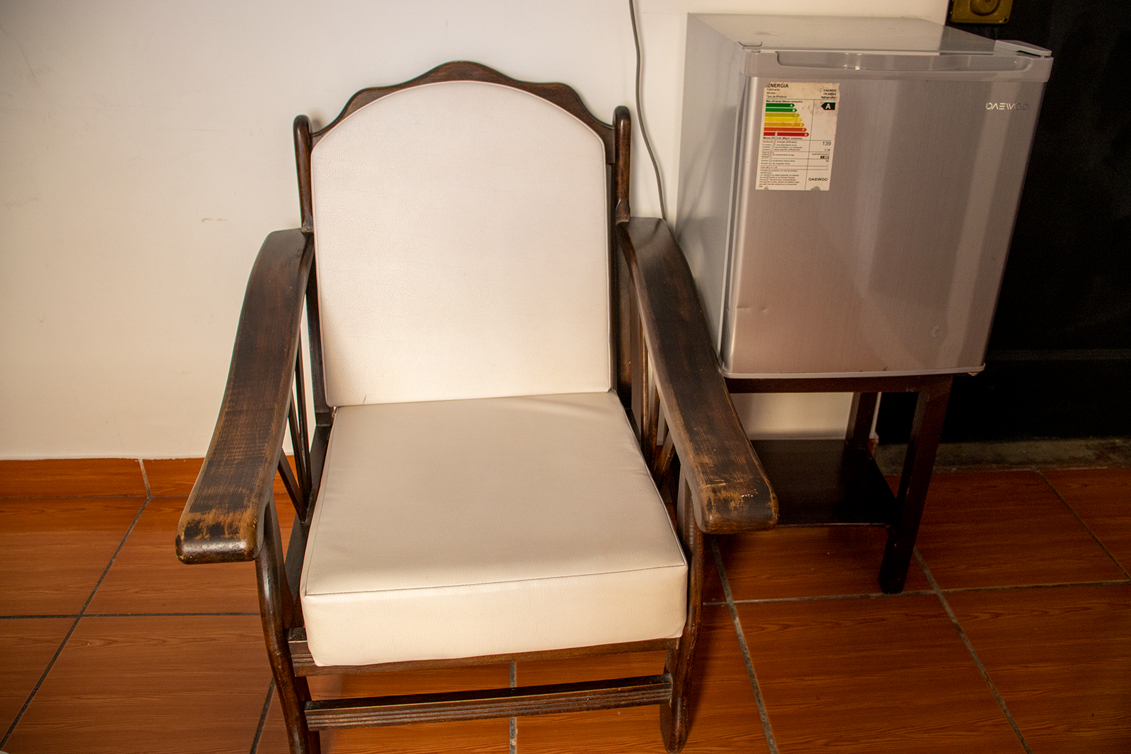 Wooden armchair with white cushions next to a small silver refrigerator on a stand.