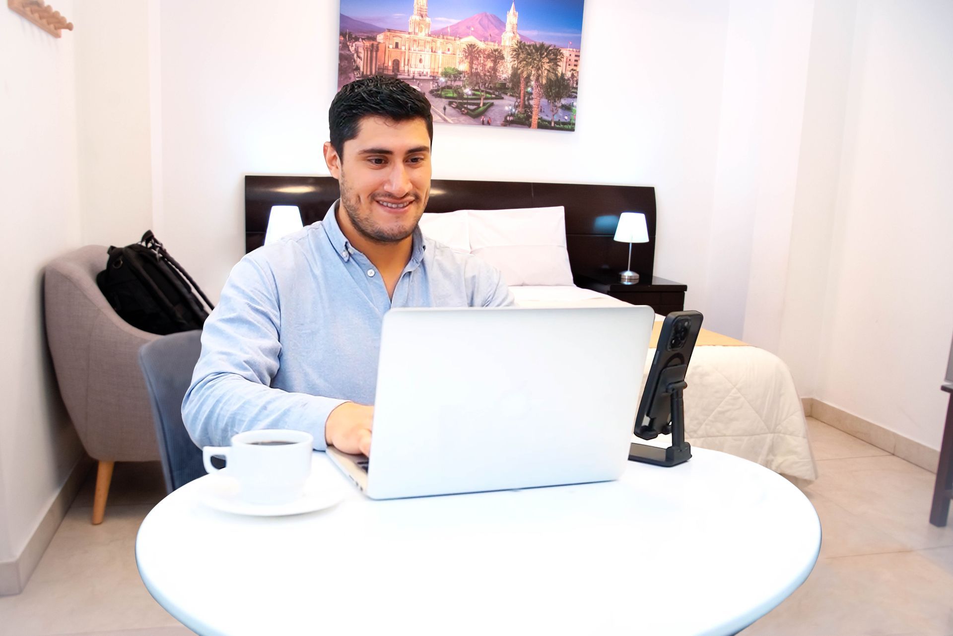 Hombre sonriendo, trabajando en laptop sobre mesa blanca en habitación de hotel con cama y arte al fondo.