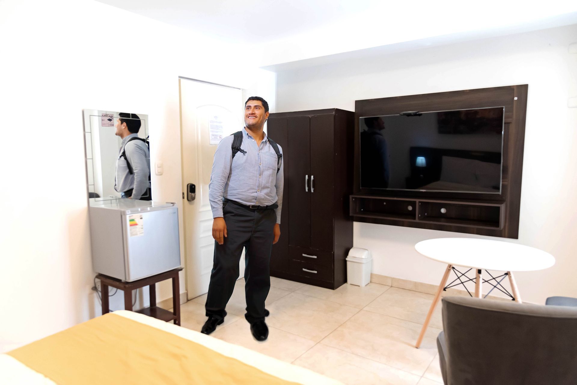 Hombre en una habitación de hotel, de pie junto a la puerta. La habitación tiene televisión, armario, nevera, mesa y silla.