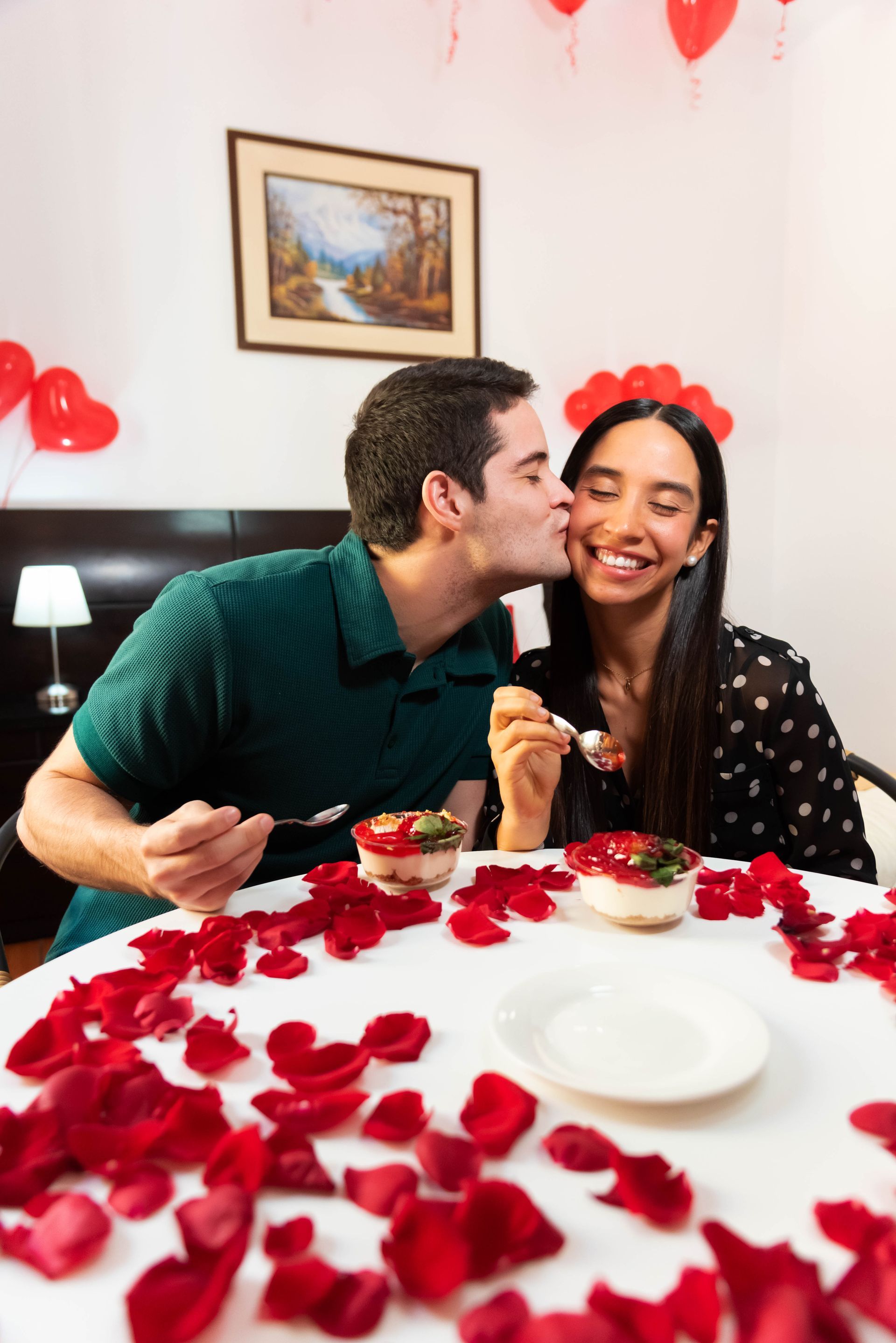 Pareja compartiendo postre, besándose, rodeadas de pétalos de rosa y globos, en una habitación decorada.