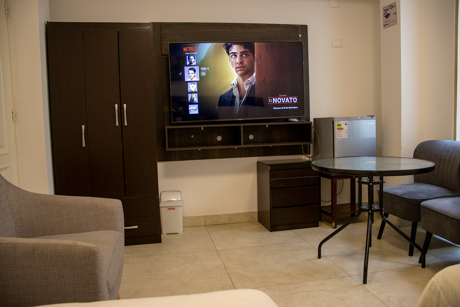 A room with a mounted TV, cabinet, mini-fridge, table, chairs and a sofa.