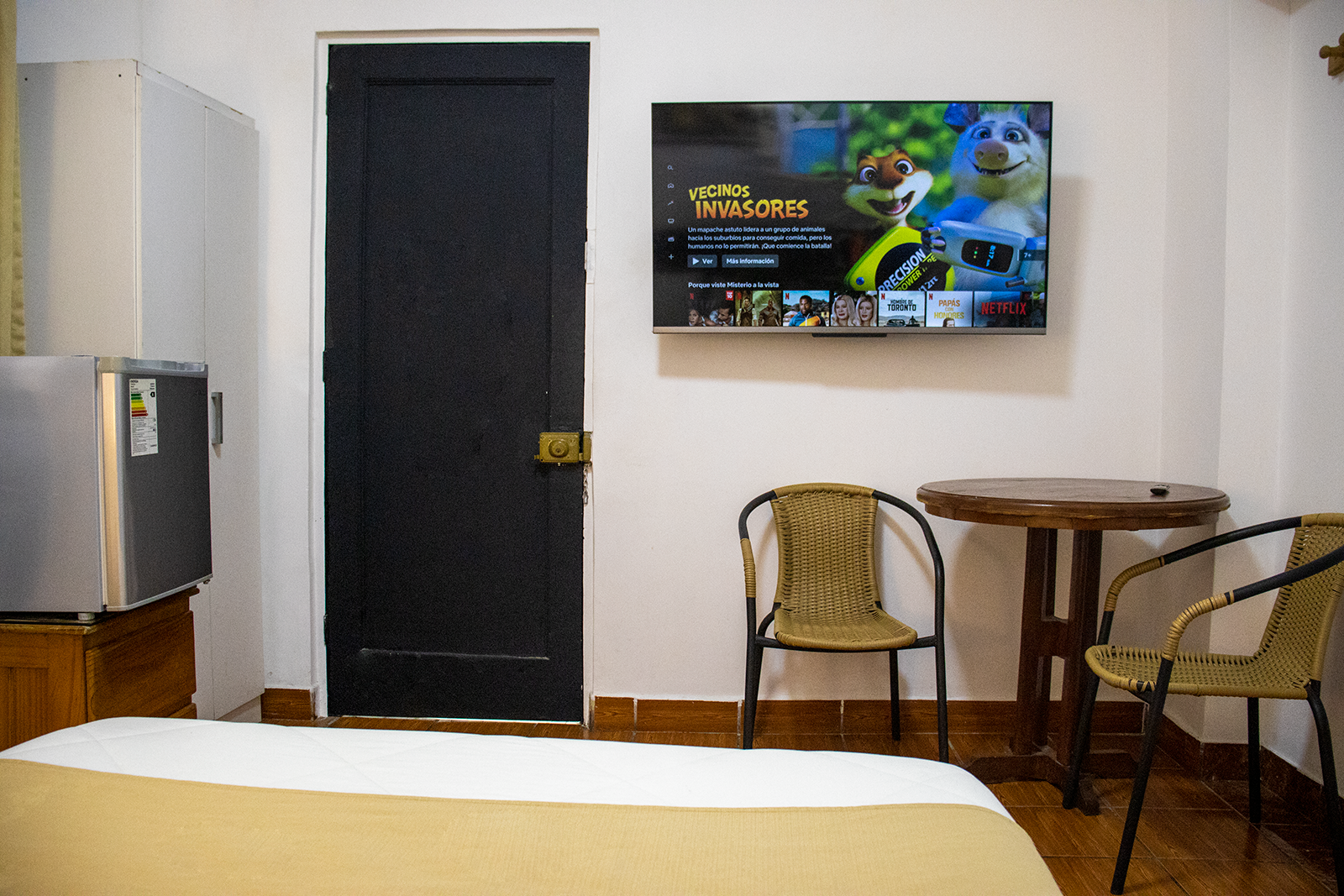 A small room with a bed, TV, chairs, a small table, fridge, and a black door.