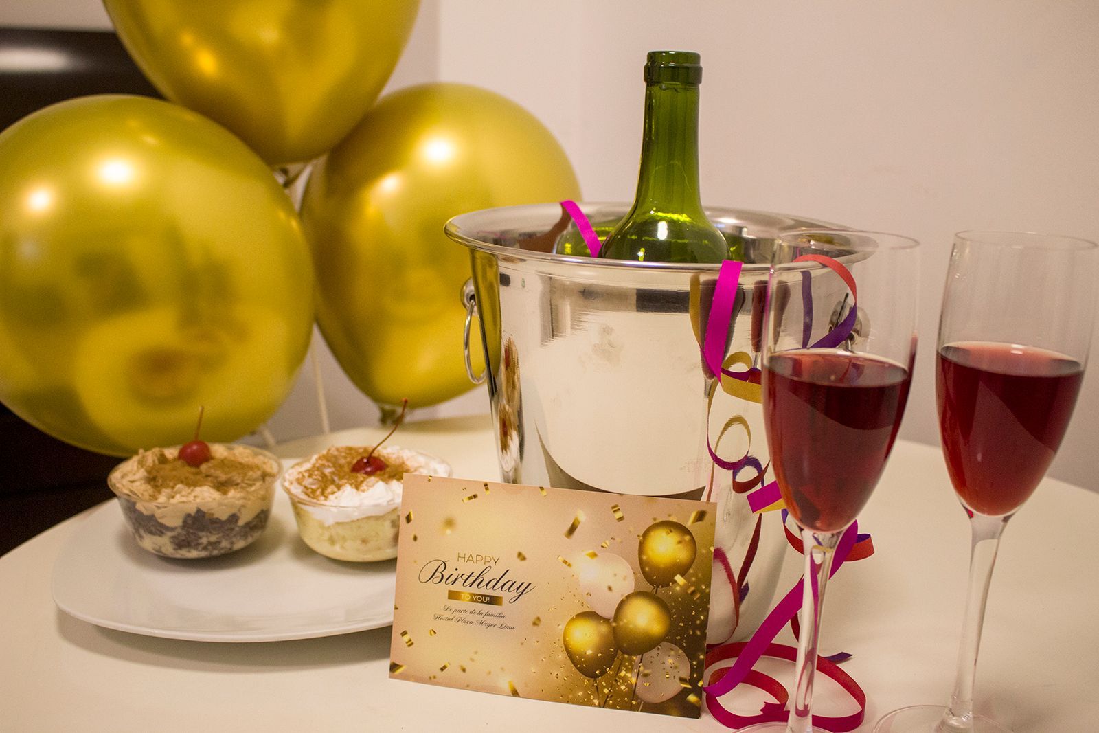 Birthday celebration: gold balloons, wine, champagne bucket, cupcakes, card.
