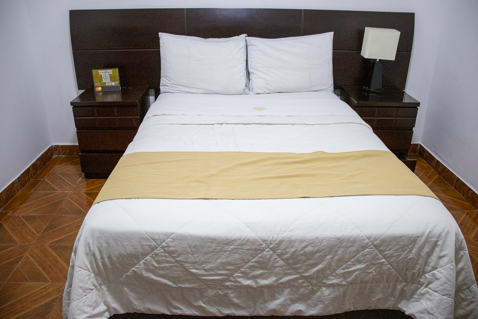 Dormitorio con cama hecha, mesitas de noche y cabecero. Sábanas blancas con tapete dorado.
