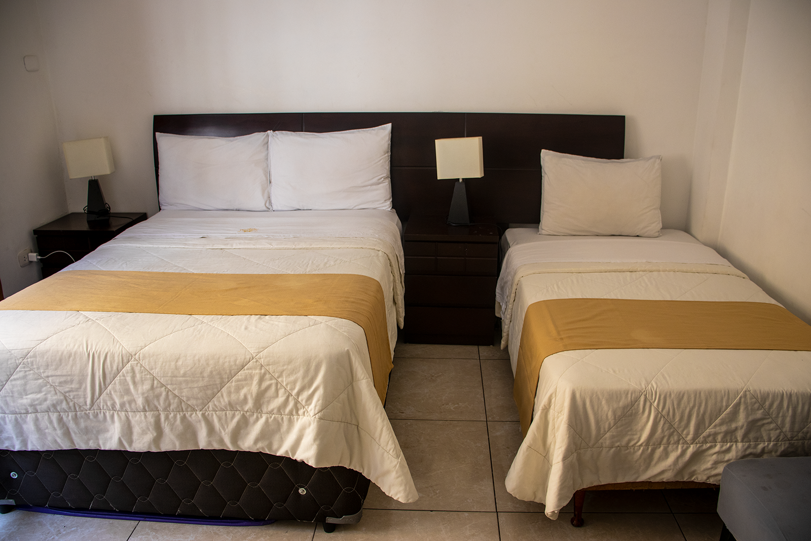 Habitación con dos camas: una doble y una individual. Ropa de cama blanca con detalles amarillos y cabecero de madera oscura.