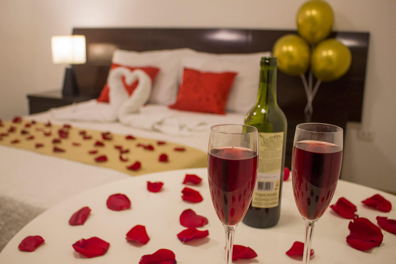 Romantic scene: bed with heart-shaped towel, wine glasses, rose petals, and balloons.