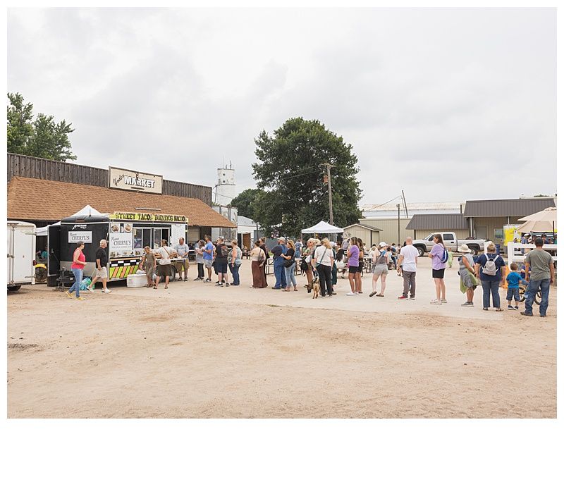 People lined up in a dusty market area beside small stalls under an overcast sky.