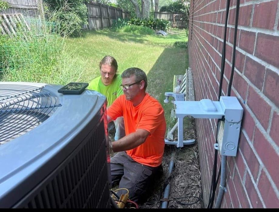 Two workers in orange and neon shirts inspect an outdoor air conditioner beside a brick house.