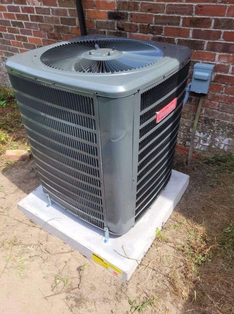Outdoor air conditioner unit beside a brick wall, sitting on a white pad in a yard